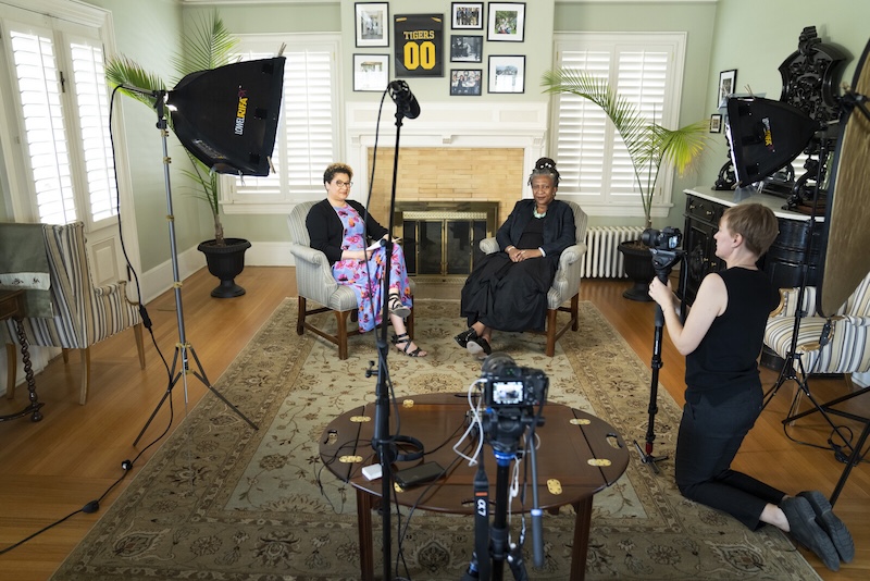 A woman kneels behind a camera recording two women in armchairs who are being interviewed
