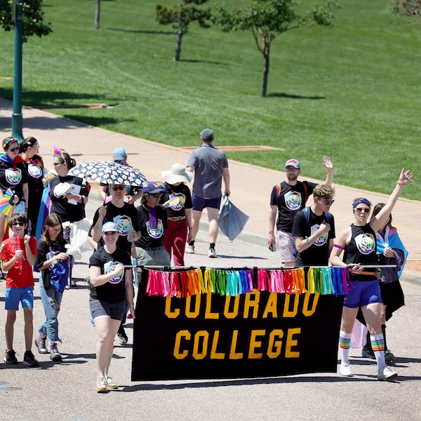 A group of people wearing rainbow versions of the CC athletics logo march carrying a banner that says Colorado College with rainbow fringe