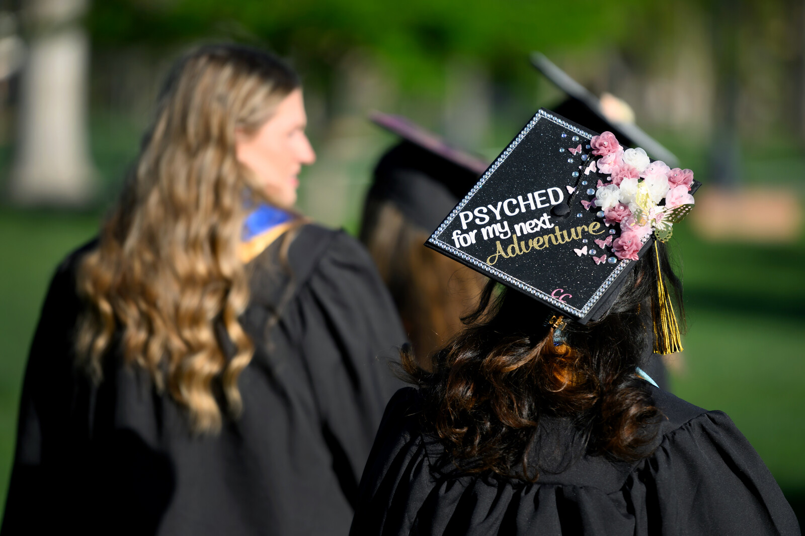 The back of two graduates walking across campus with writing on the top of one of their motorboards